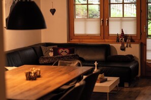 Un salon chaleureux avec un canapé en cuir noir, une table à manger en bois et une fenêtre équipée de stores.