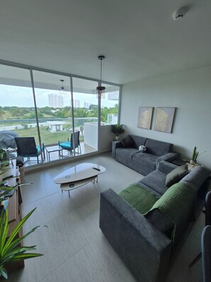 A modern living room with a grey sofa, a coffee table, and a large window offering a view of greenery and buildings. - Luxury Beach Condo in Playa Blanca | Saltwater Lagoon Access | Sleeps 6 (Río Hato)