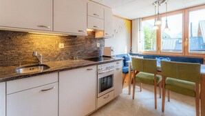 Cocina moderna con encimera de granito, pileta de acero inoxidable y muebles blancos. Incluye horno empotrado y una zona de comedor con sofá azul y sillas verdes.