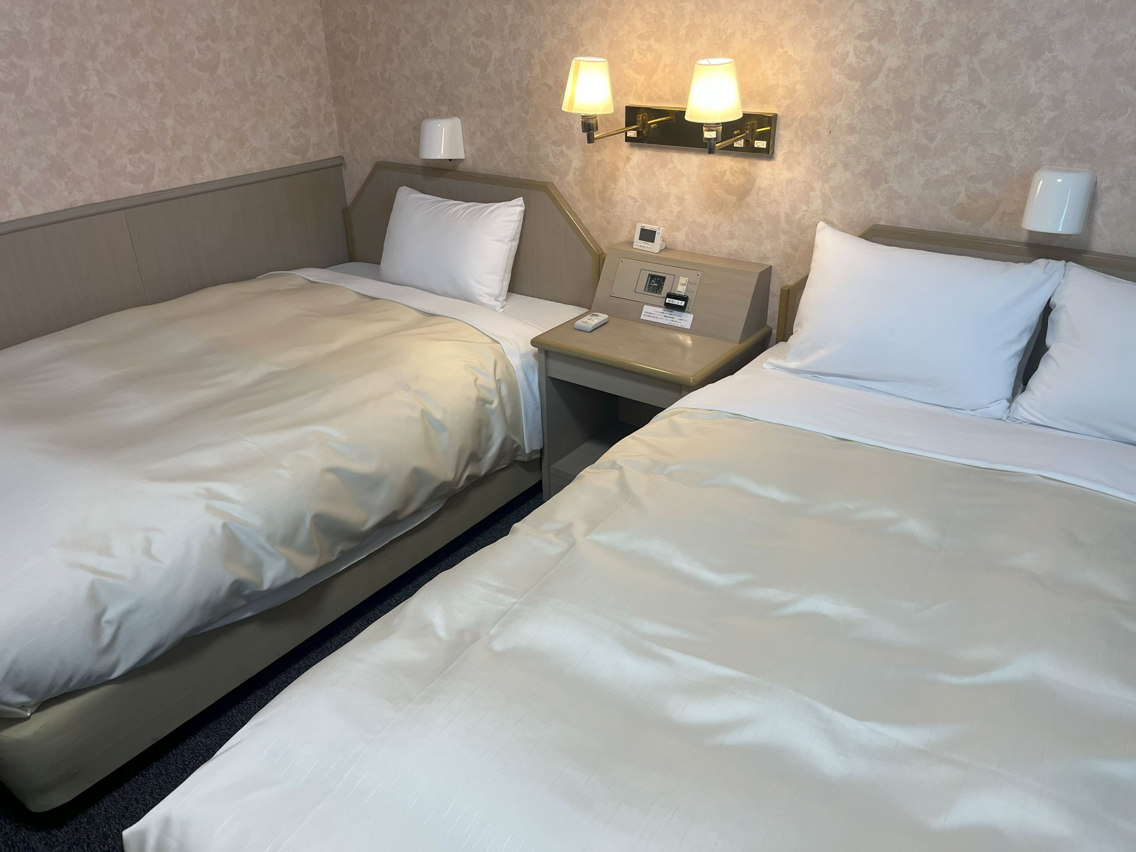 A hotel room with two beds, a nightstand with a lamp, and a wall-mounted light fixture.
