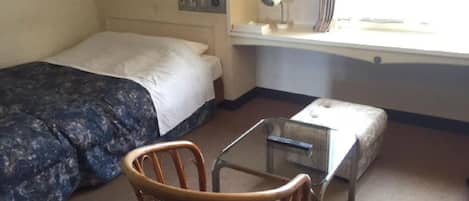 Quarto de hotel com cama, uma cadeira, uma mesa pequena, escrivaninha e janela com cortinas.