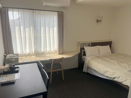 A hotel room with a bed, a desk, a chair, a window with curtains, and an air conditioning unit.