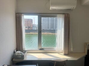 Una habitación con ventana que da a una masa de agua y edificios, un escritorio con una cesta y una tetera, y un equipo de aire acondicionado en la pared.