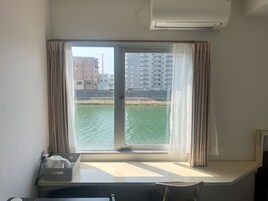A room with a window overlooking a body of water and buildings, a desk with a basket and a kettle, and an air conditioning unit on the wall.