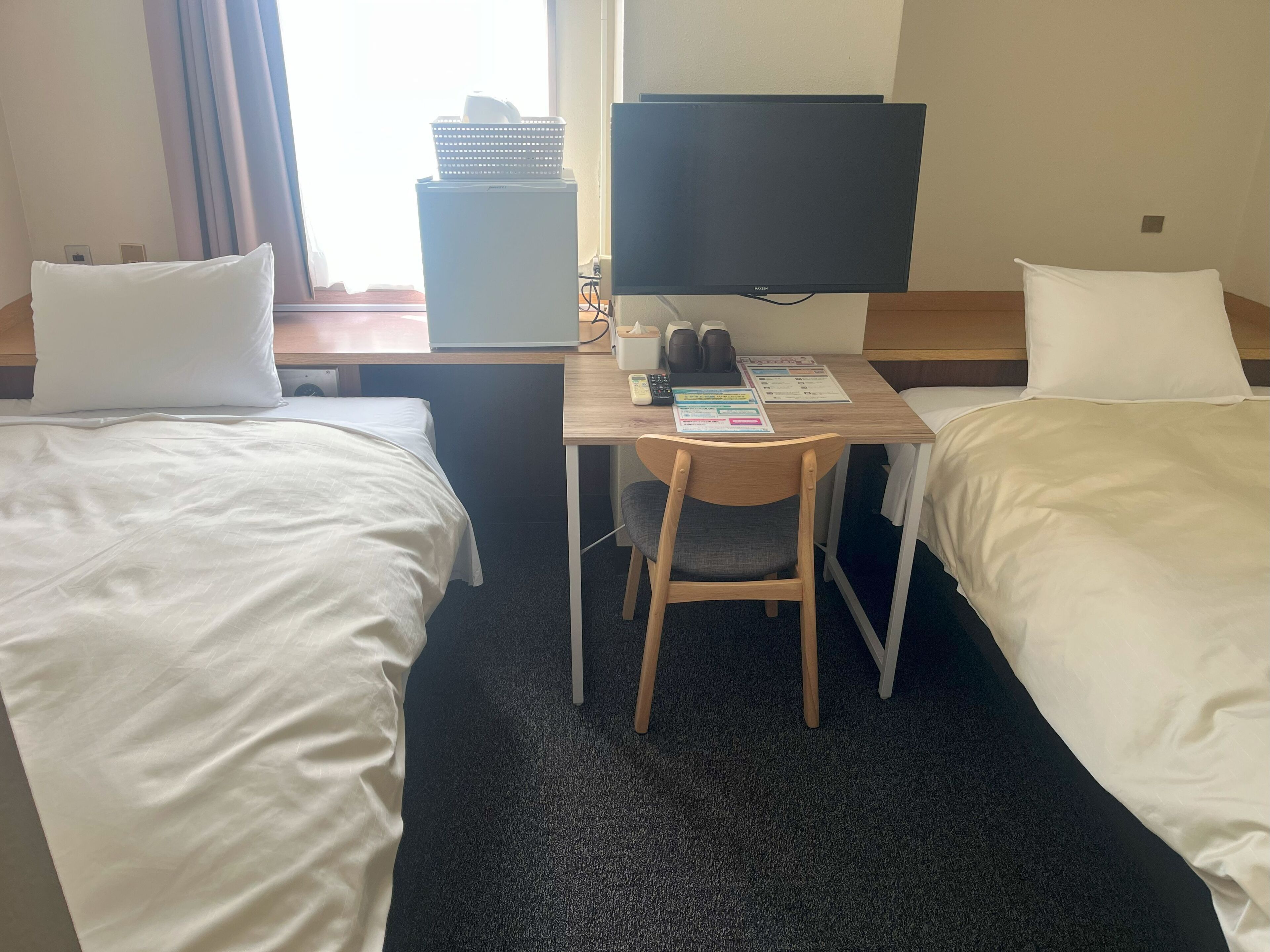 A hotel room with two single beds, a desk with a chair, a television, and a window with curtains.