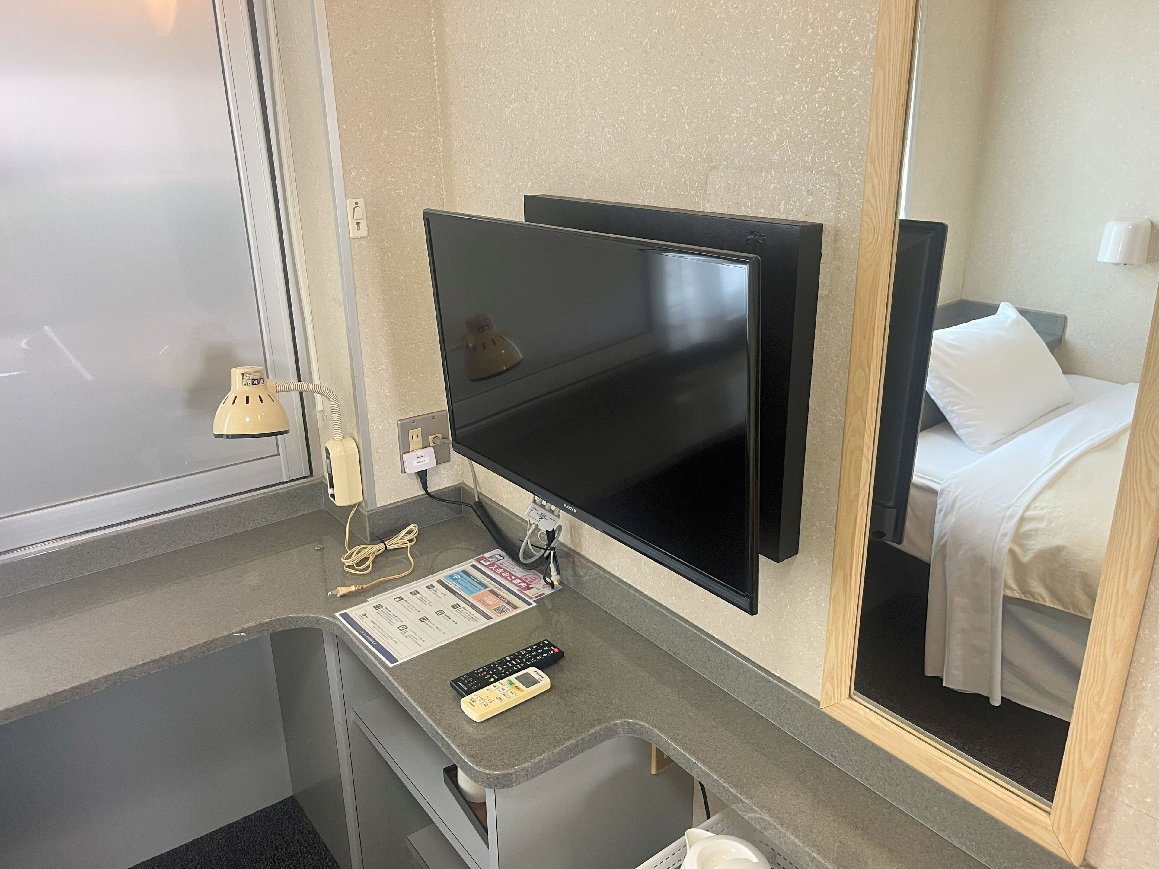 A hotel room with a flat-screen TV mounted on the wall, a small desk with a remote control and a menu card, a mirror reflecting the bed and a white pillow, and a window with a frosted glass door.