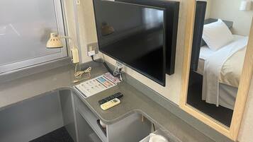 A hotel room with a flat-screen TV mounted on the wall, a small desk with a remote control and a menu card, a mirror reflecting the bed and a white pillow, and a window with a frosted glass door.