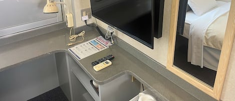 Habitación de hotel con televisor de pantalla plana montado en la pared, un escritorio pequeño con un control remoto y una tarjeta de menú, un espejo que refleja la cama y una almohada blanca, y una ventana con puerta de vidrio esmerilado.