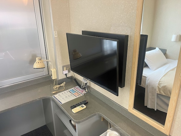 A hotel room with a flat-screen TV mounted on the wall, a small desk with a remote control and a menu card, a mirror reflecting the bed and a white pillow, and a window with a frosted glass door.