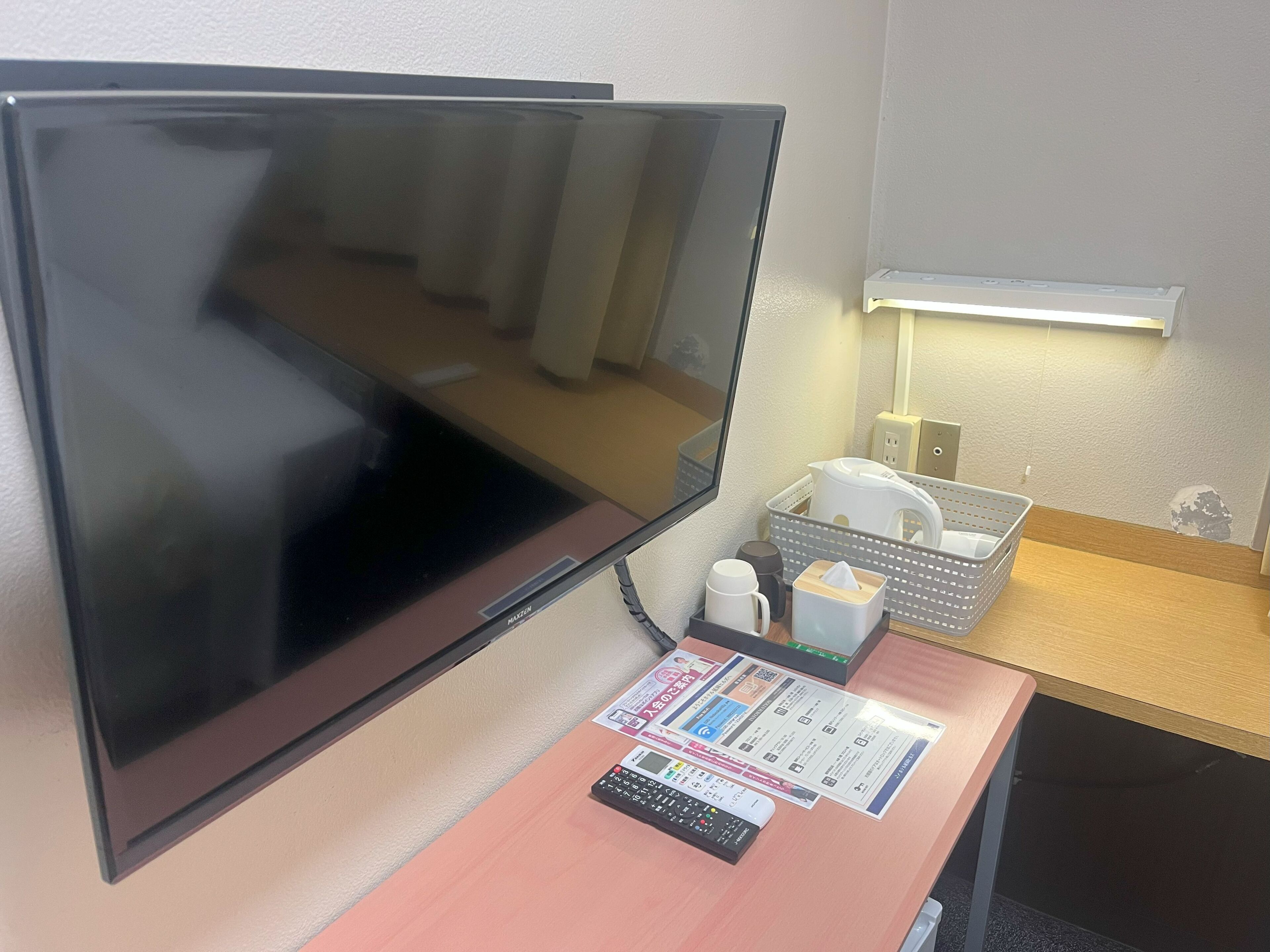A hotel room with a large flat-screen TV, a desk with a calculator, a tissue box, and a basket with a kettle and cups.