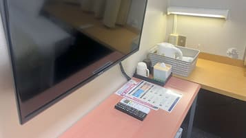 A hotel room with a large flat-screen TV, a desk with a calculator, a tissue box, and a basket with a kettle and cups.