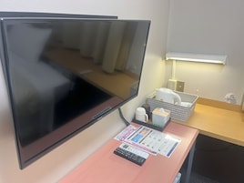 A hotel room with a large flat-screen TV, a desk with a calculator, a tissue box, and a basket with a kettle and cups.