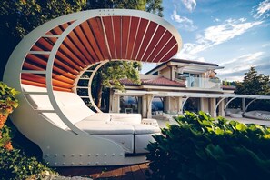 A modern outdoor seating area with a curved white structure and wooden slats, surrounded by lush greenery.
