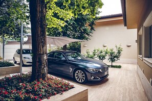 A dark-colored car parked in a driveway with a tree and flower bed in the foreground.