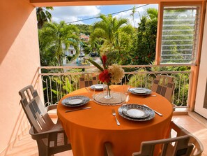 A round table with a tablecloth, plates, cutlery, and a vase with flowers, set on a balcony with a view of greenery and buildings. - Cozy apartment 150 meters from the beach (Les Trois-Îlets)