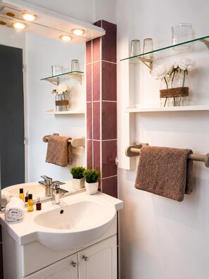 Une salle de bain moderne avec un lavabo blanc, un miroir et des étagères en verre où sont rangés les articles de toilette et des fleurs.