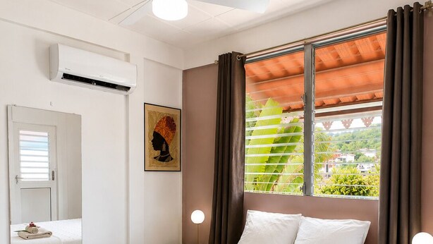Et hotelværelse med seng, ventilator, et vindue med gardiner, et natbord med lampe og et spejl.