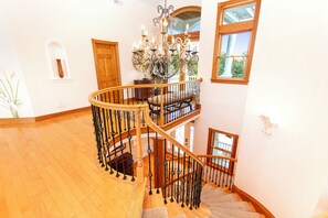 A grand staircase with a wooden handrail and black balusters, leading to a landing with a chandelier and a view of greenery outside. - Soundside-ELEV, Priv pool, HTub, Rec Rm-CC328, Pipers Perch (Corolla)