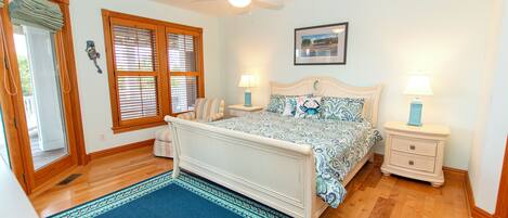 A bedroom with a bed, two bedside tables, a ceiling fan, a window with blinds, a rug, and a framed picture on the wall.