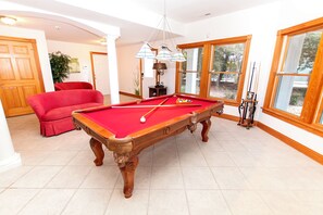 Ein Billardtisch mit roter Spielbahn, ein rotes Sofa und ein Fenster mit Blick auf Bäume.