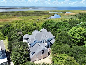 A large house with multiple levels and a complex roof structure, surrounded by dense greenery and a water body in the background. - Soundside-ELEV, Priv pool, HTub, Rec Rm-CC328, Pipers Perch (Corolla)