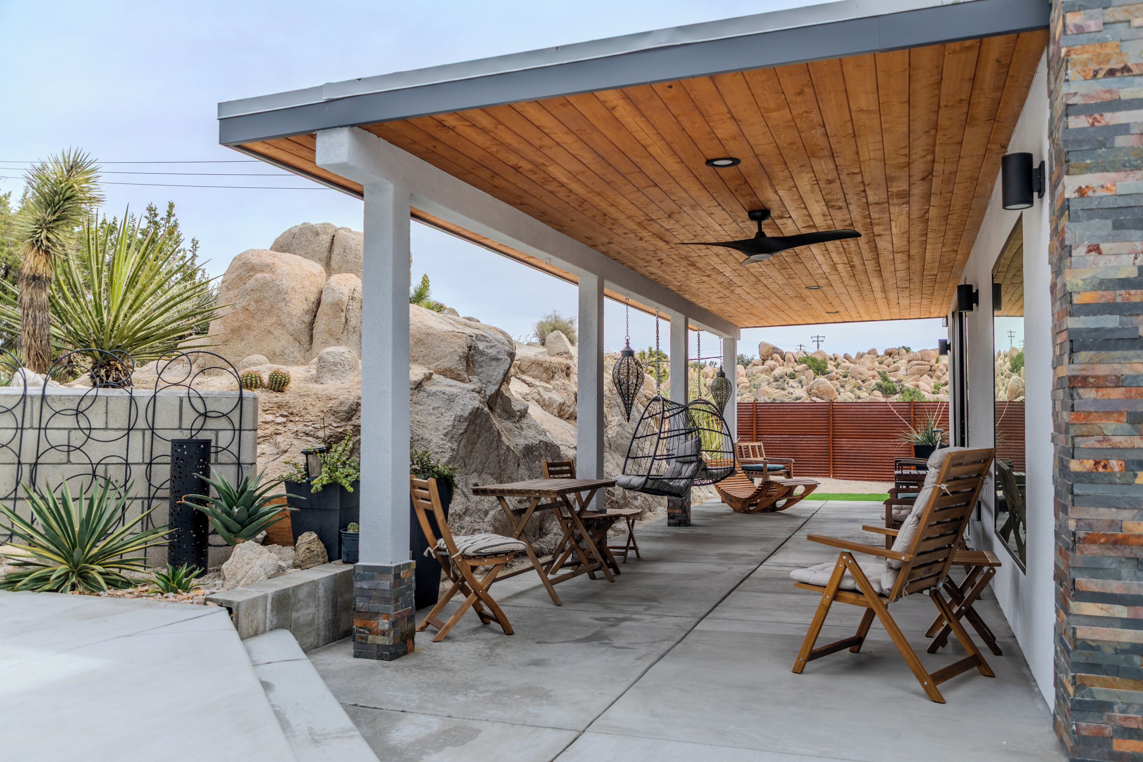 An outdoor patio area with a wooden ceiling, stone walls, and a seating arrangement including chairs and tables.