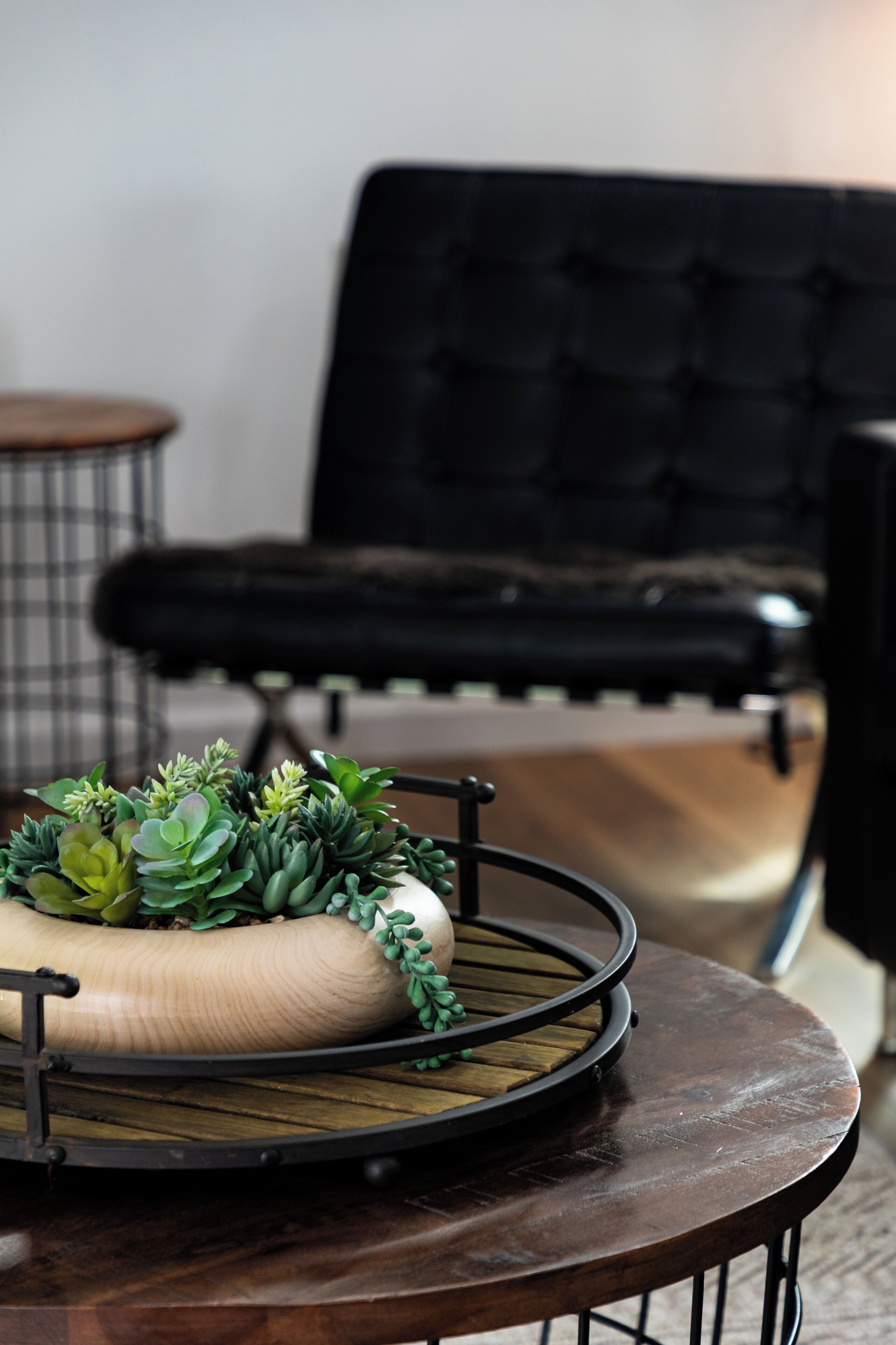 A round wooden table with a metal tray holding green plants, a black upholstered chair, and a plaid-patterned stool in a room with a neutral color scheme.