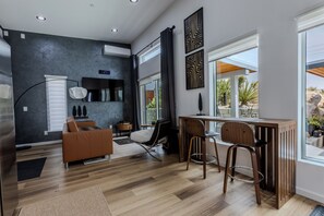Ein modernes Wohnzimmer mit dunkler Wand, Holzboden, einem braunen Sofa, einem Esstisch mit zwei Stühlen und einem großen Fenster mit Blick ins Freie.