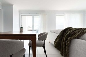 A modern living room with a sofa, a dining table, and chairs. There is a large window with curtains, and a view of the outdoors. - Wake Up to Stunning Tamar Valley Views (Riverside)