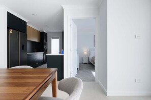 A modern kitchen with dark cabinetry, a wooden dining table, and a view into a bedroom with a bed and nightstand. - Wake Up to Stunning Tamar Valley Views (Riverside)