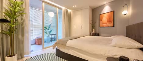 A modern bedroom with a large bed, a sliding glass door to a balcony, a potted plant, and a striped rug.