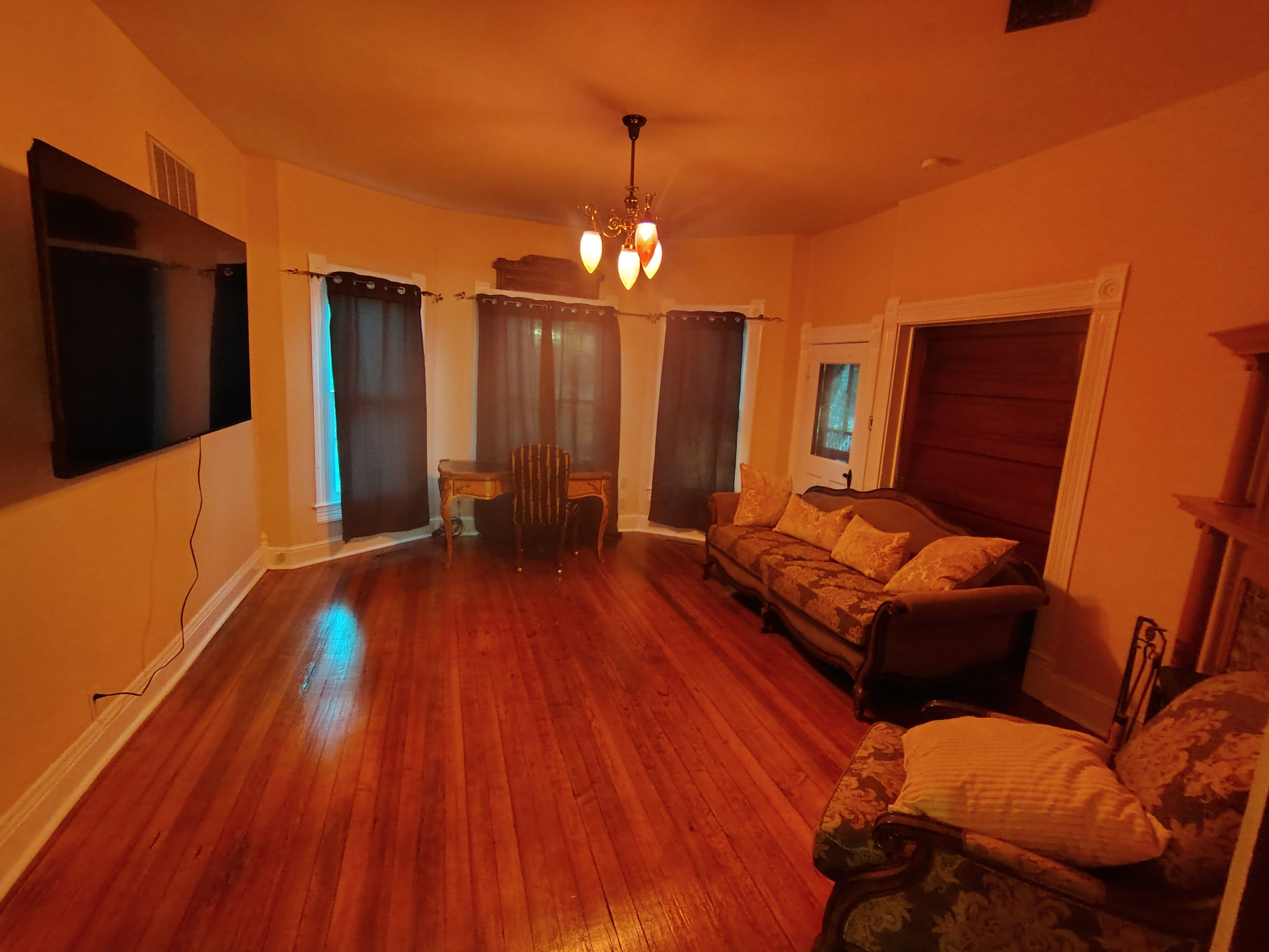 A living room with a wooden floor, a sofa, a chair, a television mounted on the wall, and a chandelier.