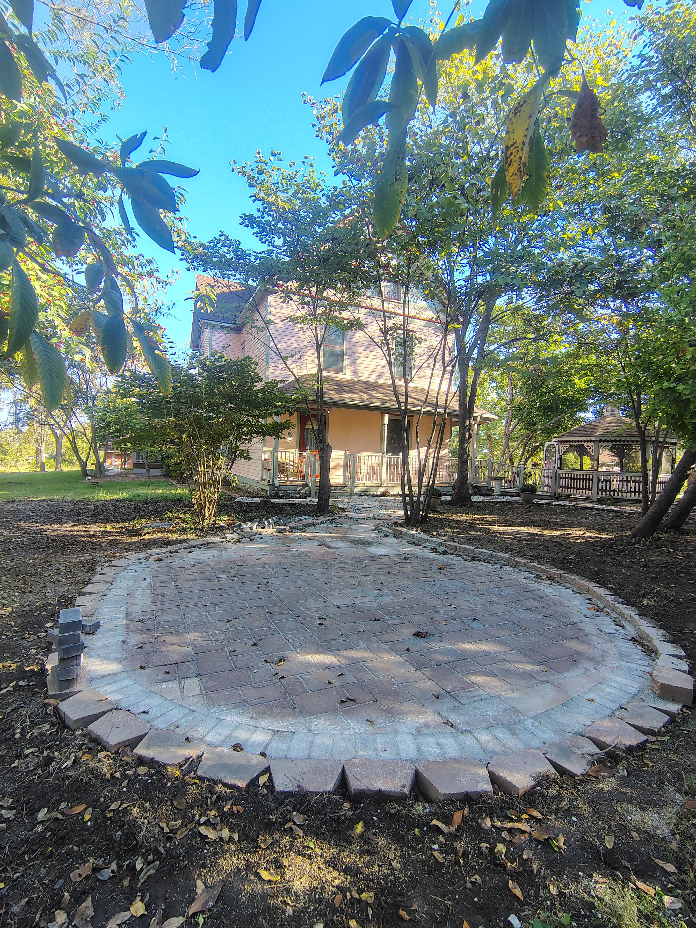 Um pátio pavimentado com um gazebo, rodeado por árvores e uma casa.