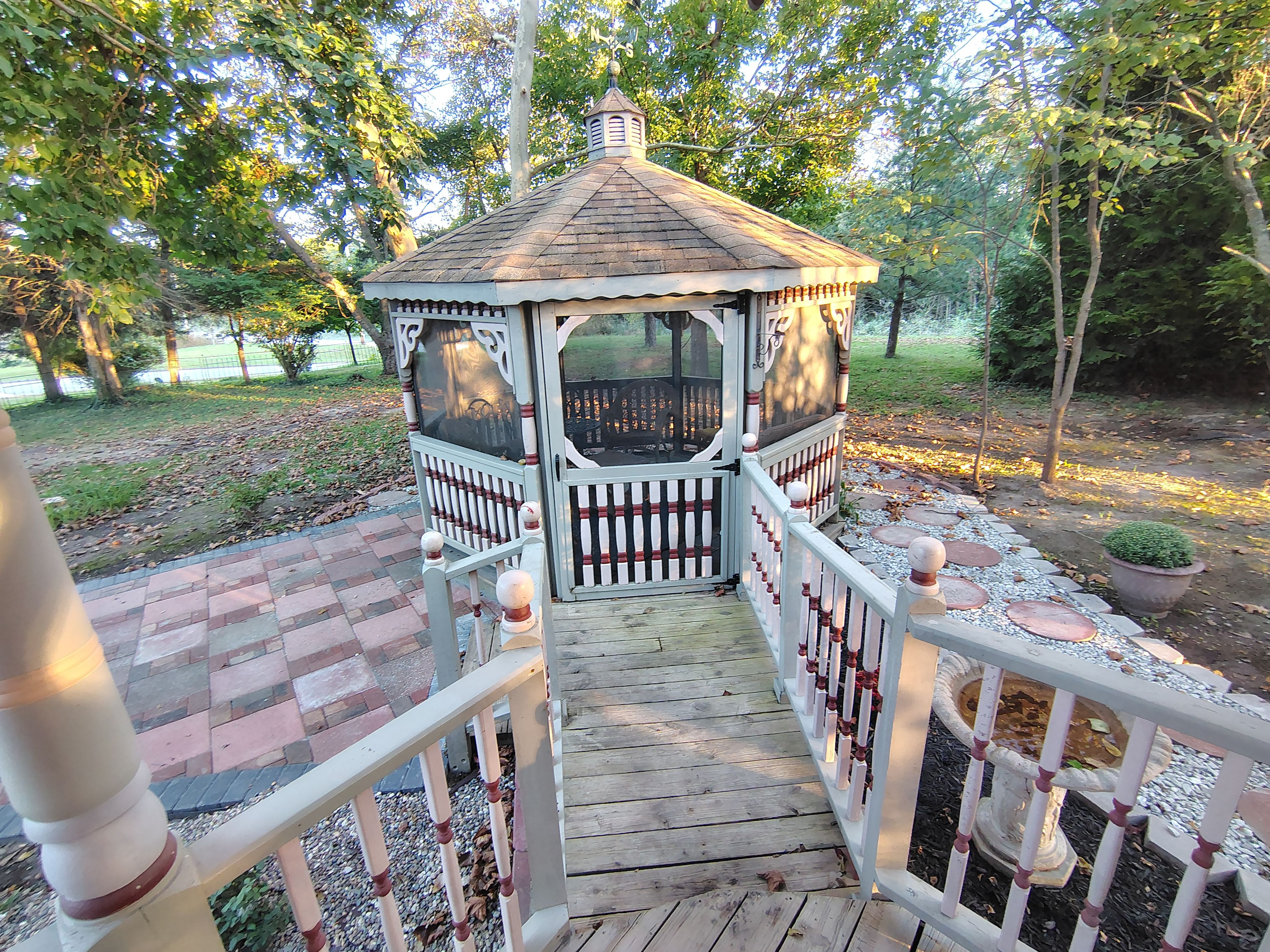Um gazebo com deck de madeira e grades brancas, rodeado por árvores e um caminho de tijolos.