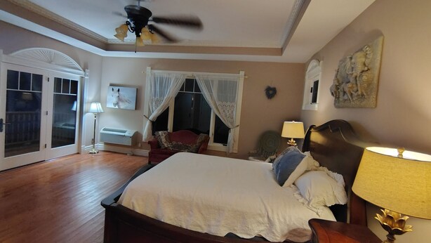 A bedroom with a large bed, a ceiling fan, a window with curtains, a wall-mounted artwork, and a nightstand with a lamp.