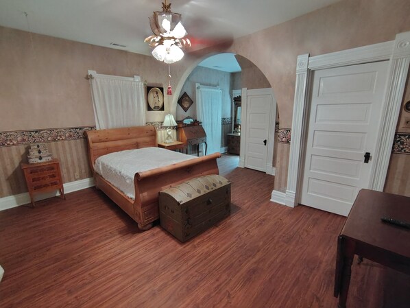 Un dormitorio con una cama de madera, una mesita de noche, un aparador y un cómoda. Hay una puerta que da a otra habitación.