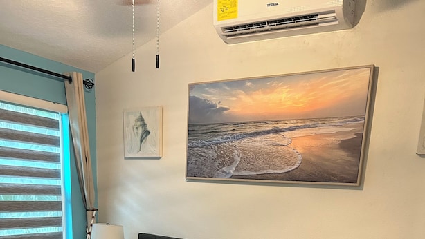 Ein ordentlich bezogenes Bett mit einem aufgerollten gelben Handtuch, ein Strandbild und eine kleine Kommode mit dekorativer Front.