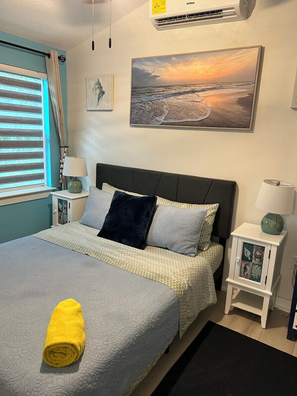 A neatly made bed with a rolled yellow towel, a painting of a beach scene, and a small cabinet with a decorative front.
