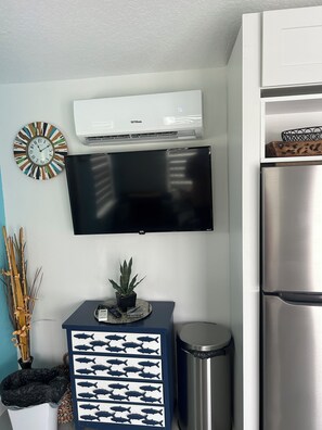 A kitchen with a flat-screen TV mounted above a black cabinet, a stainless steel refrigerator, and a wall-mounted air conditioner.