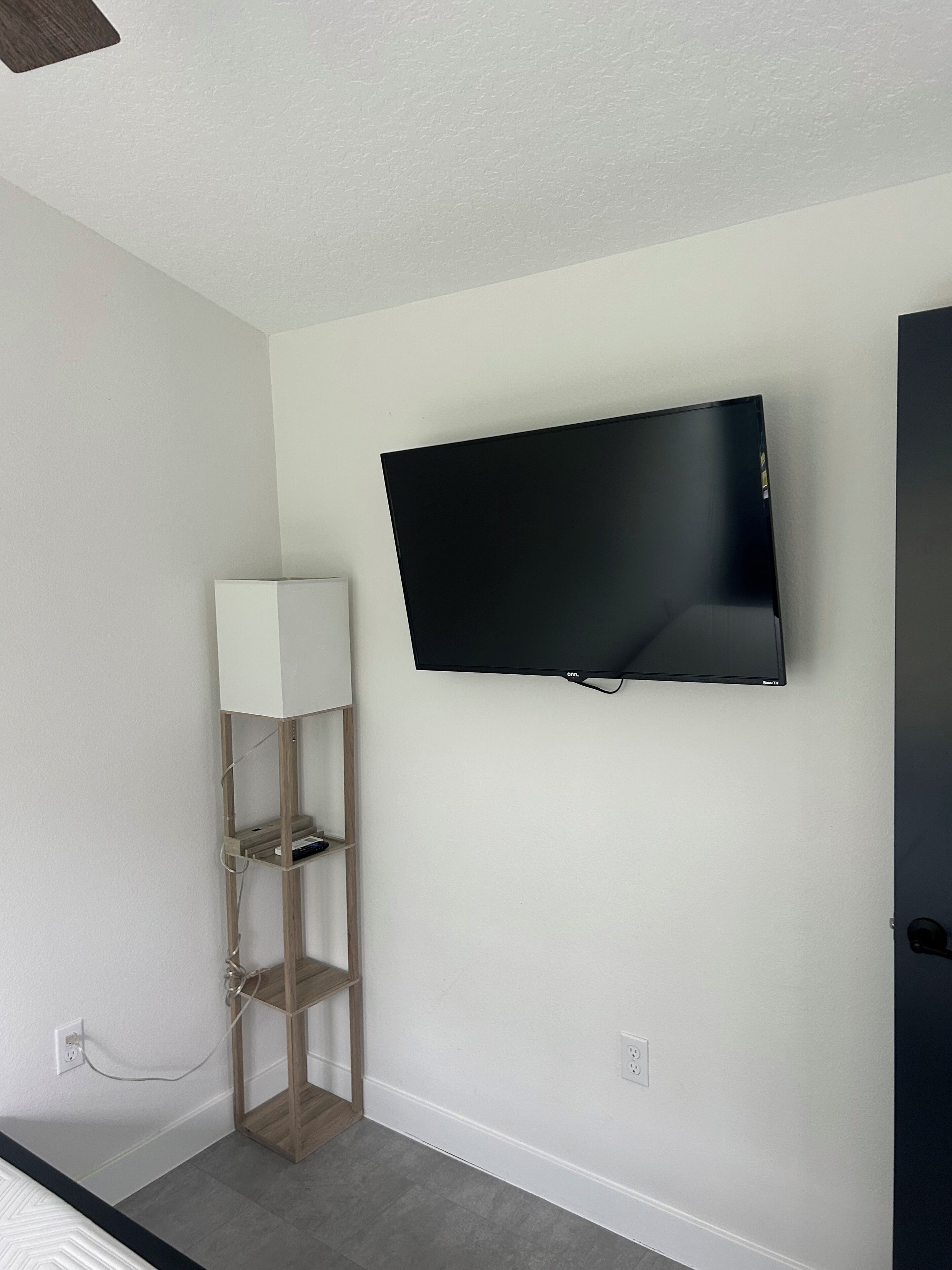 Una habitación con una televisión de pantalla plana montada, una lámpara de pie de madera y una pared blanca.