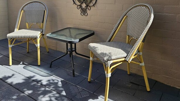 Two wicker chairs with metal frames and a small glass table, positioned against a brick wall with a decorative metal piece above. - Garden Retreat on Orr – Your Peaceful Hideaway (Shepparton)