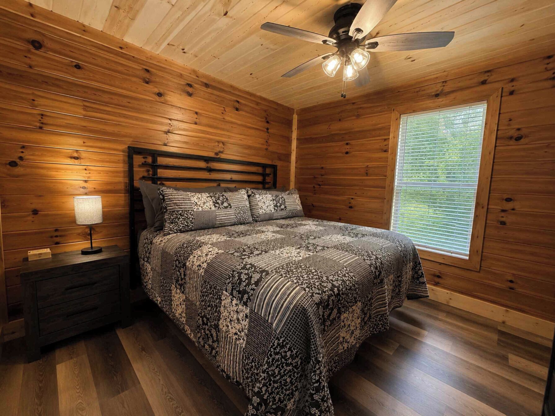 Un dormitorio con techo de madera, una cama con ropa de cama estampada, una mesita de noche con lámpara y una ventana con persianas.