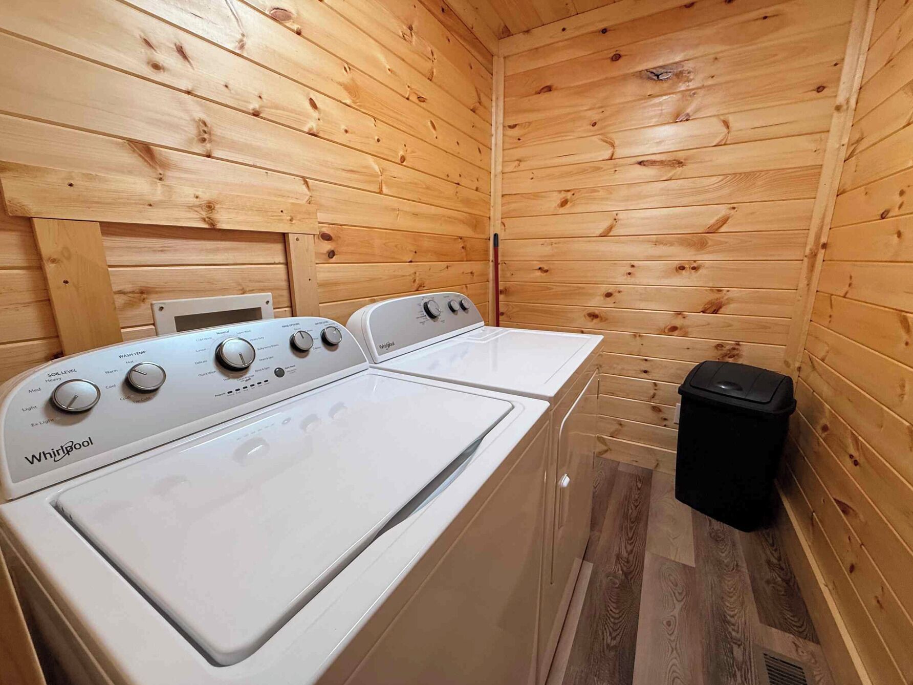 Un lavadero con dos lavadoras Whirlpool, paredes de madera y un bote de basura negro.