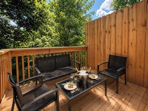 An outdoor deck with a wooden railing, two black metal chairs, a glass table with a metal pot, and a bottle of champagne.