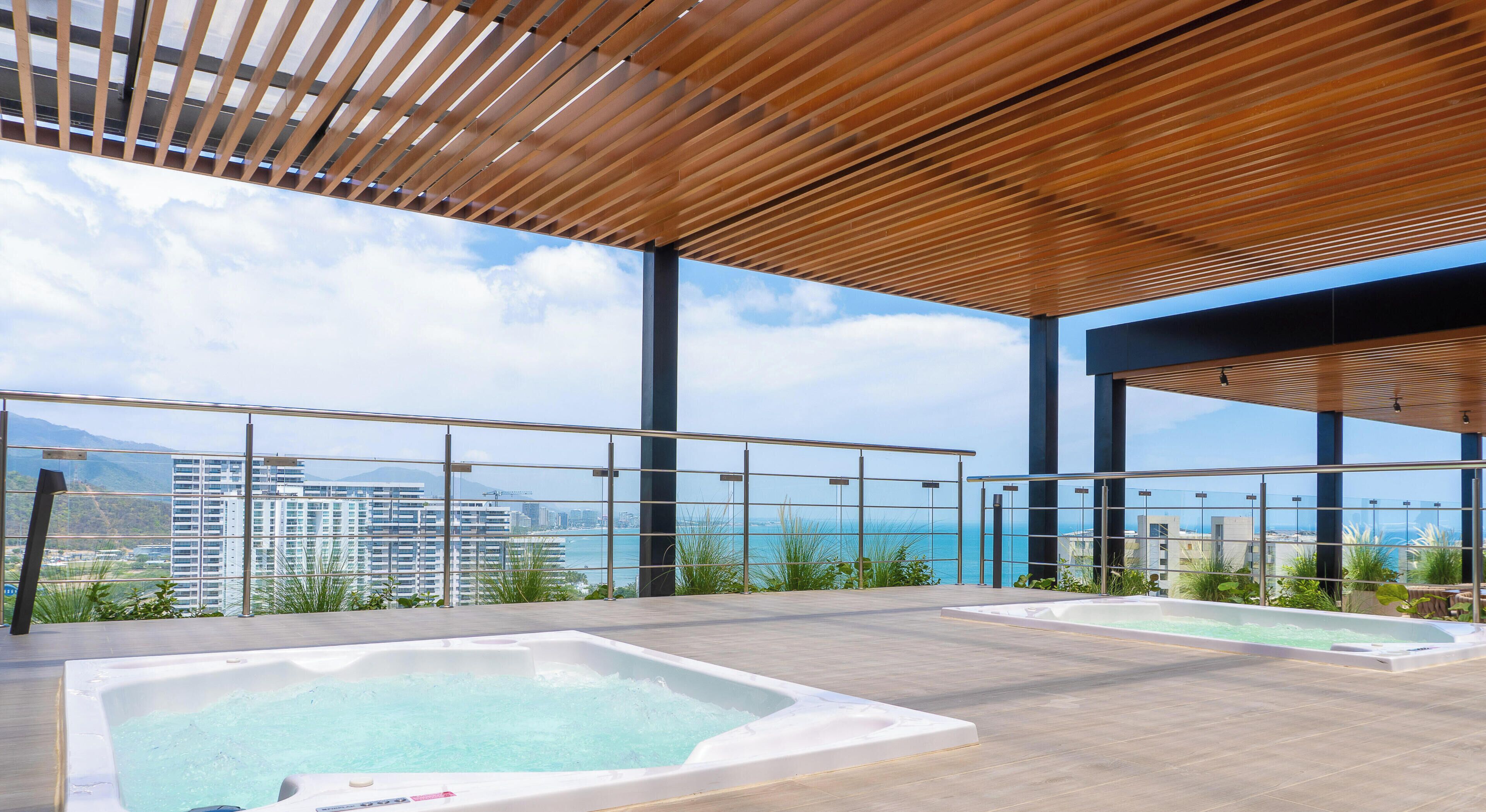 A rooftop terrace with two hot tubs, a wooden ceiling, and a view of the ocean and city skyline.
