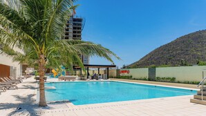 A swimming pool with lounge chairs and a palm tree, surrounded by a building and a hilly landscape.