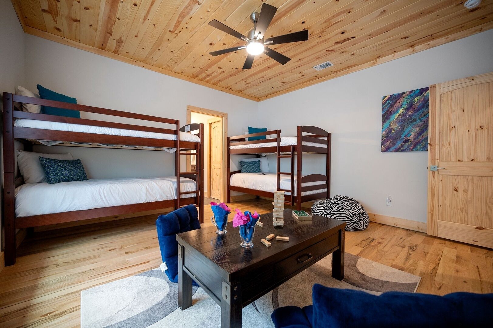A room with bunk beds, a ceiling fan, a table with a vase and blocks, and a zebra-patterned bean bag.
