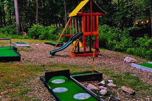 A playground with a slide and a swing set, surrounded by trees and pebbles. - Fall Promo! Modern • Mini Golf • Hot Tub • Hot Tub (Broken Bow)