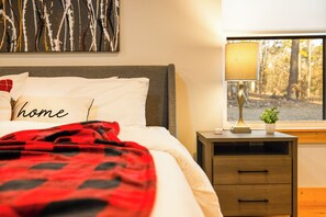 A neatly made bed with a red and black blanket, a pillow with the word "home", a lamp on a bedside table, and a window with a view of trees. - Fall Promo! Modern • Mini Golf • Hot Tub • Hot Tub (Broken Bow)