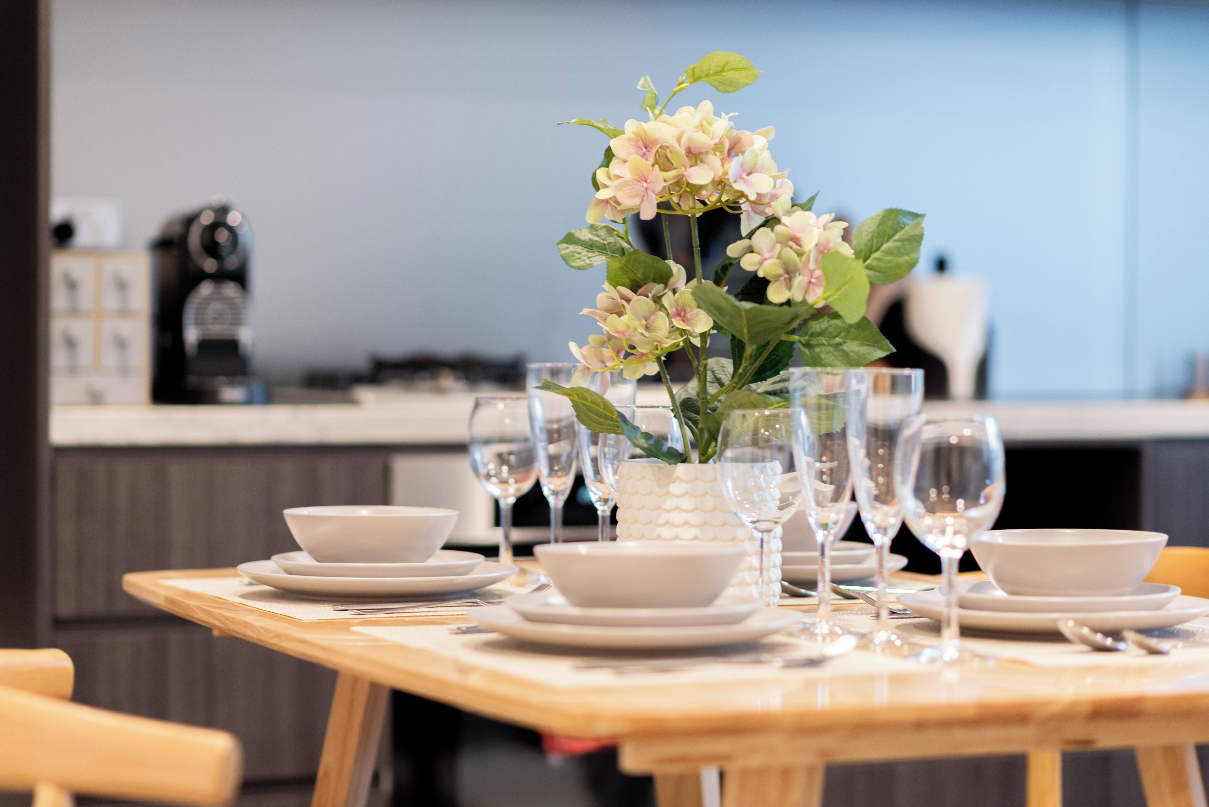 A dining table set with white plates, bowls, and wine glasses, with a vase of flowers in the center.
